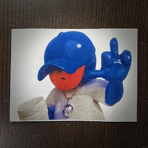 May include: A cartoon figure wearing a blue baseball cap and a white shirt, giving the middle finger. The figure has a red face and a silver chain with a pendant. The image is on a white background.