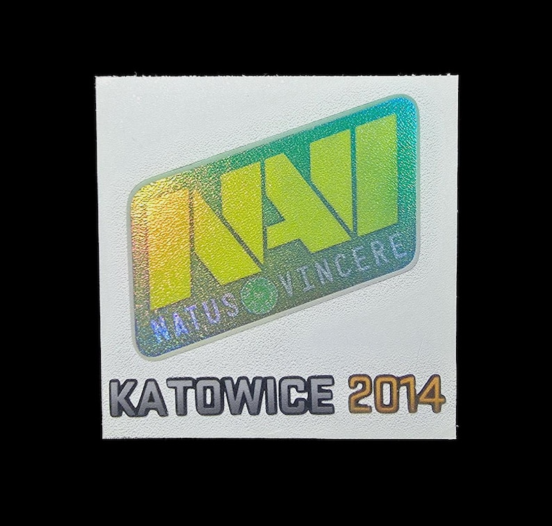 May include: A square white sticker featuring a holographic NaVi logo with the text Natus Vincere in a slanted rectangular frame. Below the logo, the text KATOWICE 2014 is printed in bold, dark gray capital letters on the white background surface.