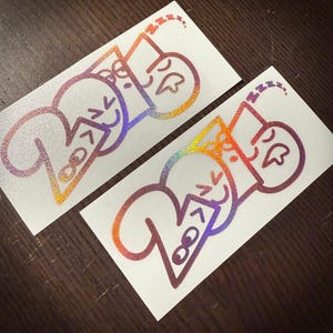 May include: Two stickers with a holographic design. Each sticker features the word "Zzz" with a sleeping face and a smiling face. The stickers have a gradient of colours, including orange, purple, and blue.