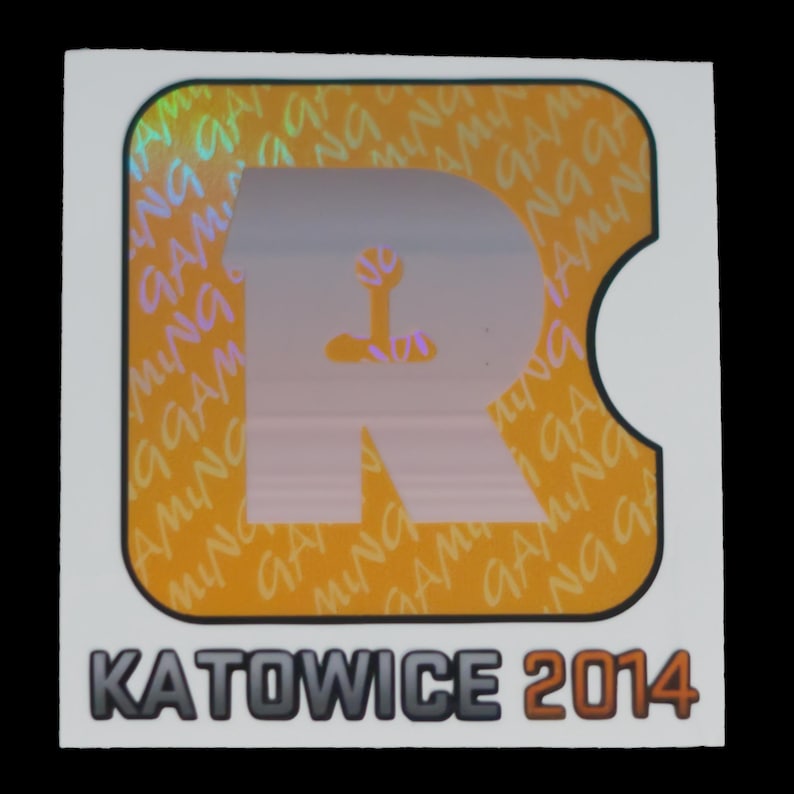 May include: A square sticker featuring a large, light-colored letter R centered on an orange background with a repeating holographic pattern of the word GAMING. Below the orange square, the text KATOWICE 2014 is printed in bold, dark letters.