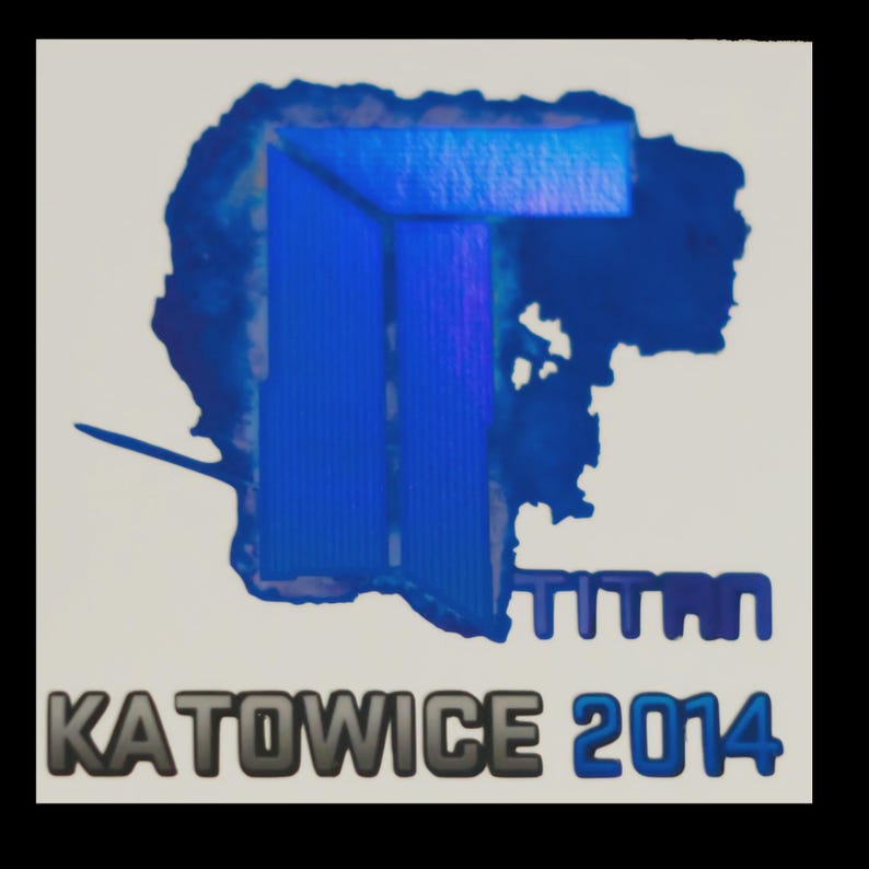 May include: A square sticker featuring a large, blue, holographic-style letter T logo with a jagged, ink-blot-like outline. Below the logo, the word TITAN is printed in blue, followed by the text KATOWICE 2014 in bold, dark gray and blue lettering.