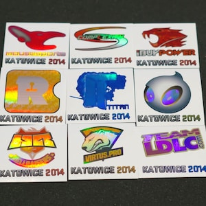 May include: A collection of nine square stickers featuring various esports team logos from the Katowice 2014 tournament. Each sticker displays a unique team emblem with holographic or metallic finishes and the text Katowice 2014 printed below.
