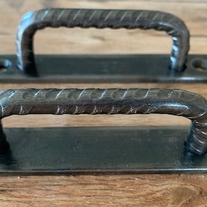 Peut inclure: Deux poignées de porte en métal foncé avec une poignée texturée en forme de barre d'armature. Chaque poignée a une base rectangulaire avec des trous pré-percés pour le montage. La quincaillerie est présentée sur une surface en bois.