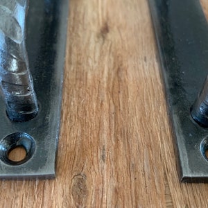 May include: Two black, hand-forged metal door pulls. Each pull has a rectangular base with a curved handle. The handles have a textured, hammered appearance. The pulls are mounted on a light brown wooden surface.