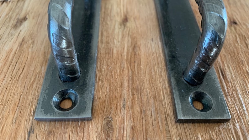 May include: Two black, hand-forged metal door pulls. Each pull has a rectangular base with a curved handle. The handles have a textured, hammered appearance. The pulls are mounted on a light brown wooden surface.