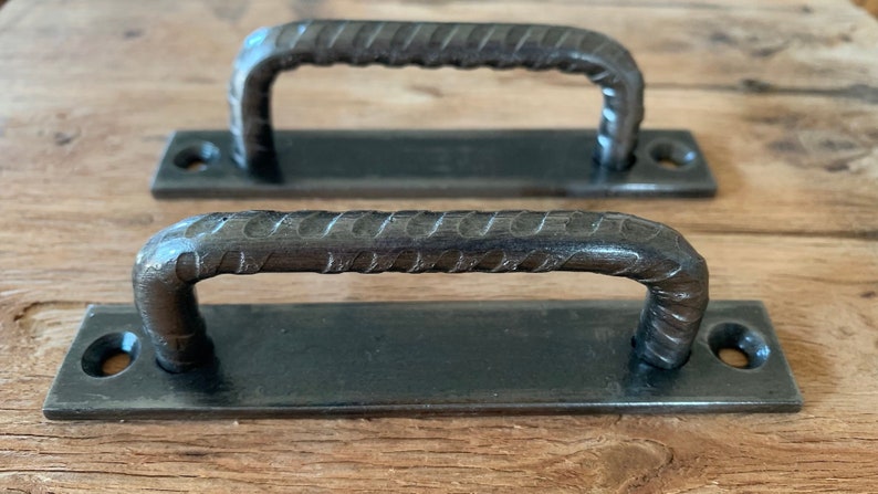 May include: Two rustic, dark metal door pulls with a textured, rebar-like handle design. Each pull has a rectangular base with pre-drilled holes for mounting. The hardware is displayed on a wooden surface.