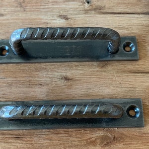 May include: Two dark gray metal handles with a textured, rebar-like design. One handle is arched, and the other is straight. Each handle is attached to a rectangular plate with two holes for mounting. The handles are likely for a cabinet or drawer.