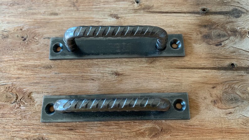 May include: Two dark gray metal handles with a textured, rebar-like design. One handle is arched, and the other is straight. Each handle is attached to a rectangular plate with two holes for mounting. The handles are likely for a cabinet or drawer.
