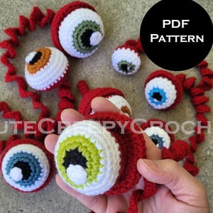 Könnte beinhalten: Gehäkelte rote und weiße Augäpfel mit verschiedenen Irisfarben, darunter Grün, Blau und Orange. Die Augen sind durch rote gehäkelte Ranken verbunden. Der Text "PDF PATTERN" befindet sich in einem schwarzen Kreis. Der Text "CUTECROCHETPYC" ist ebenfalls sichtbar.