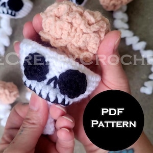 May include: Crocheted skull and brain ornaments in white, black, and peach. The skull has black eye sockets and a detailed jawline. The brain is a peach color. The image also includes the text "PDF PATTERN".