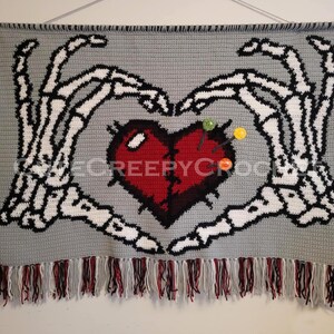 Puede incluir: Un tapiz de pared de ganchillo gris con un coraz&oacute;n rojo y manos de esqueleto blancas. El coraz&oacute;n tiene alfileres. El borde inferior tiene una franja de hilo rojo, negro y blanco. El texto "@CREEPY CROCHET" es visible.