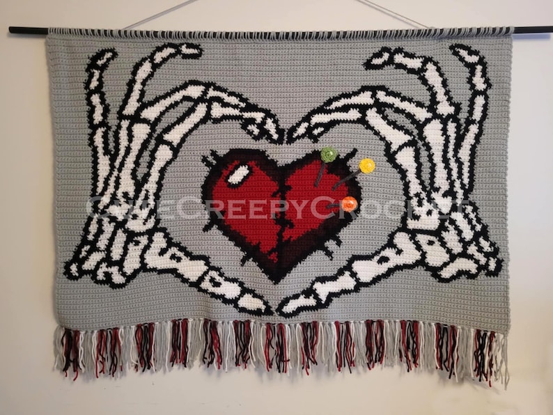 Puede incluir: Un tapiz de pared de ganchillo gris con un coraz&oacute;n rojo y manos de esqueleto blancas. El coraz&oacute;n tiene alfileres. El borde inferior tiene una franja de hilo rojo, negro y blanco. El texto "@CREEPY CROCHET" es visible.