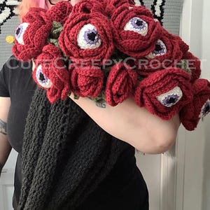 May include: A bouquet of red crochet flowers, each with a white and purple eye center. The flowers are held in a black crocheted wrap. The text "cutecreepycrochet" is visible.