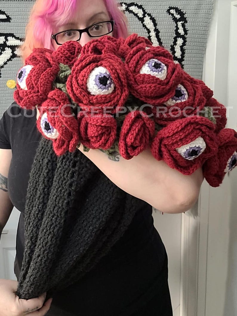 May include: A bouquet of red crochet flowers, each with a white and purple eye center. The flowers are held in a black crocheted wrap. The text "cutecreepycrochet" is visible.