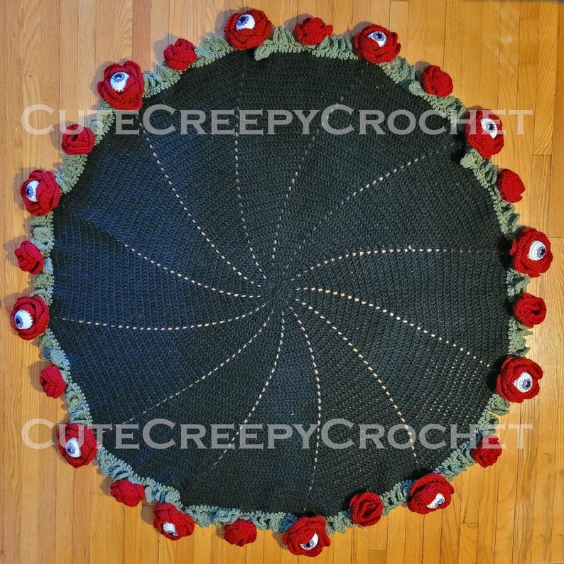May include: A round, dark green crocheted blanket with a spiral design. The edge is decorated with red roses and green leaves, each rose featuring a blue and white eyeball. The text "Cute Creepy Crochet" is visible.