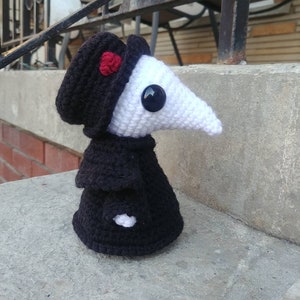 May include: A handmade crochet plague doctor doll. The doll features a white beak, a black hat with a red flower, and a black cloak. The doll has a large black button for an eye. The doll is sitting on a concrete surface.