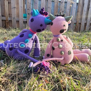 May include: Two handmade crochet monster dolls. One is purple with teal and black spots, the other is pink with green and black spots. Both have horns and are sitting on grass. The text "CUTECG" is visible on the purple doll.