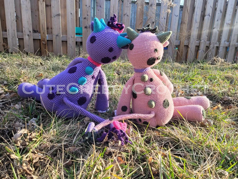 May include: Two handmade crochet monster dolls. One is purple with teal and black spots, the other is pink with green and black spots. Both have horns and are sitting on grass. The text "CUTECG" is visible on the purple doll.