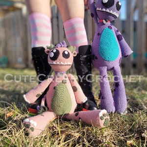 May include: Two handmade crochet monster dolls. One is pink with green accents and black spots, the other is purple with a teal belly and black spots. The dolls have large eyes and are sitting in the grass. The text "CUTECROCHET" is visible.