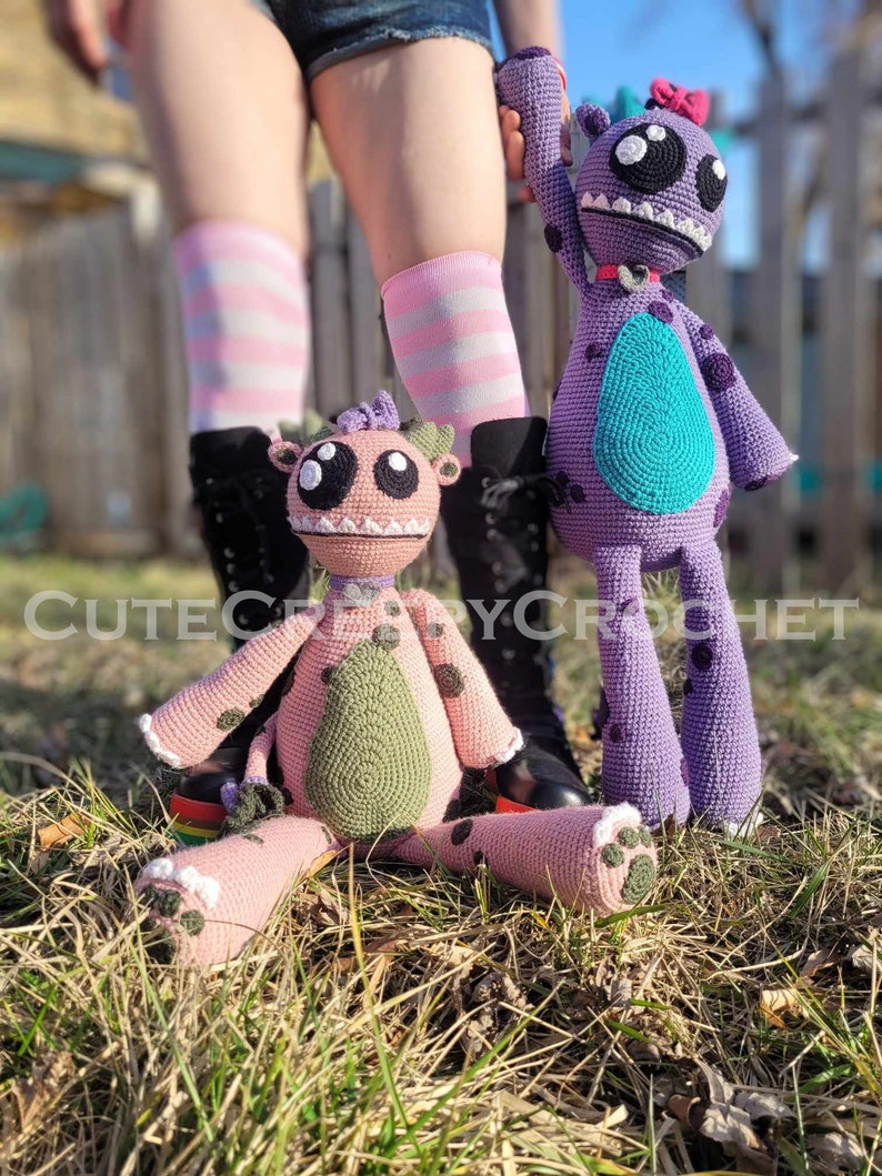 May include: Two handmade crochet monster dolls. One is pink with green accents and black spots, the other is purple with a teal belly and black spots. The dolls have large eyes and are sitting in the grass. The text "CUTECROCHET" is visible.