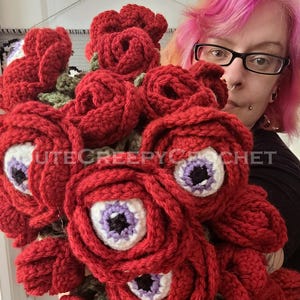 May include: A bouquet of red crochet roses with white and purple eye centers. The text "CUTECREEPYCROCHET" is visible on the image. The handmade flowers have green crochet leaves.