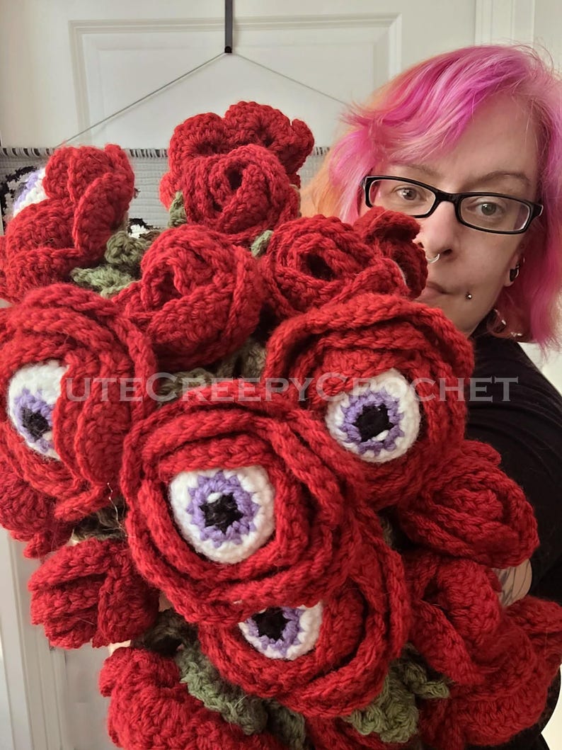 May include: A bouquet of red crochet roses with white and purple eye centers. The text "CUTECREEPYCROCHET" is visible on the image. The handmade flowers have green crochet leaves.