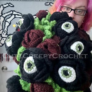 May include: A handmade crochet bouquet featuring black roses with white and green eye-shaped centers, burgundy roses, and green foliage. The bouquet is labeled with the text "ittycreepycrochet".