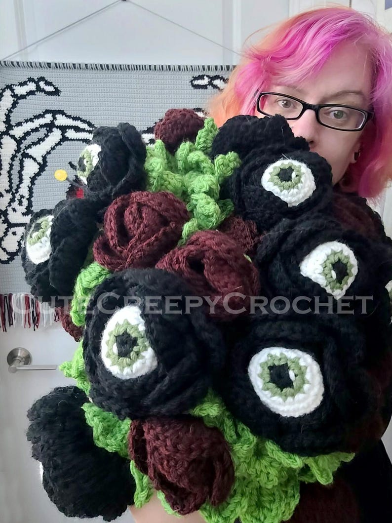 May include: A handmade crochet bouquet featuring black roses with white and green eye-shaped centers, burgundy roses, and green foliage. The bouquet is labeled with the text "ittycreepycrochet".