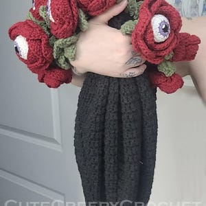 May include: A crochet bouquet of red roses with white and purple eyeball centers, green leaves, and a black stem. The text "CuteCreepyCrochet" is visible in the image. The bouquet is held in a hand.