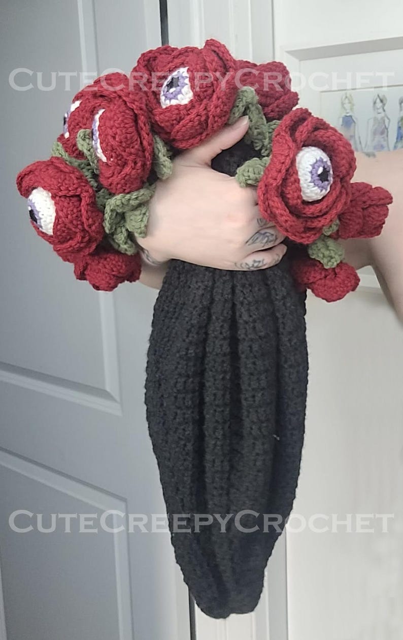 May include: A crochet bouquet of red roses with white and purple eyeball centers, green leaves, and a black stem. The text "CuteCreepyCrochet" is visible in the image. The bouquet is held in a hand.