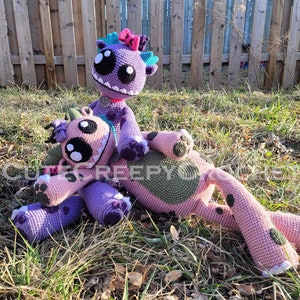May include: Two handmade crochet monster dolls in shades of pink and purple, with large eyes and toothy grins. One doll is seated on top of the other, both lying on grass. The dolls have various details, including spots, bows, and spikes.
