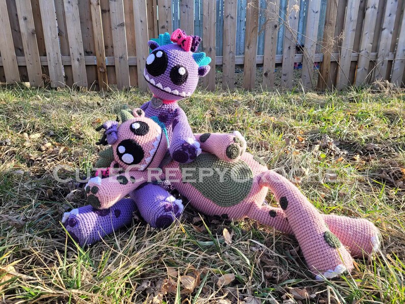 May include: Two handmade crochet monster dolls in shades of pink and purple, with large eyes and toothy grins. One doll is seated on top of the other, both lying on grass. The dolls have various details, including spots, bows, and spikes.