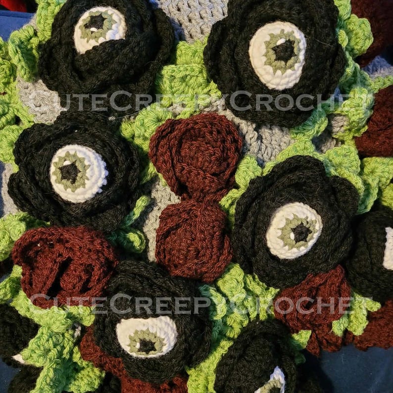 May include: Close-up of a crocheted blanket featuring large, black flower-like shapes with white and green centers, and smaller burgundy and green accents. The text "CUTECREEPYCROCHE" is visible. The blanket has a unique, handmade aesthetic.