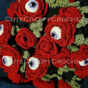May include: A crocheted bouquet of red roses with white and purple eye centers. The green leaves and dark background create a striking contrast. The text "CUTECREEPYCROCHET" is visible on the image. This unique handmade item is perfect for Halloween or gothic decor.