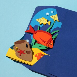 May include: A blue paper envelope with a yellow, green, and red paper cutout design inside. The design features a red fish, two yellow fish, a brown clay pot, and green seaweed. The design is a representation of an underwater scene.