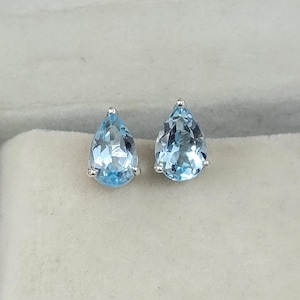May include: A pair of light blue teardrop-shaped gemstone stud earrings. The gemstones are set in a silver metal. The earrings are displayed on a light gray surface.