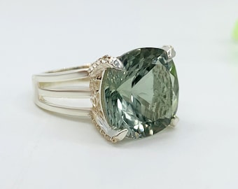 May include: A silver ring featuring a large, square-cut, pale green gemstone. The ring has a split shank design with three parallel bands and ornate detailing around the gemstone setting. The gemstone is faceted, reflecting light.