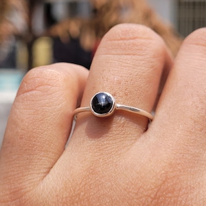 Puede incluir: Primer plano de un anillo de plata con una pequeña gema negra redonda. El anillo es sencillo y elegante, con una banda fina. La gema está engastada en un bisel y el anillo se lleva en un dedo.