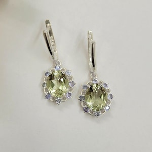 May include: A pair of silver earrings featuring oval, pale green gemstones encircled by small, light blue gemstones. The earrings have a lever back closure.