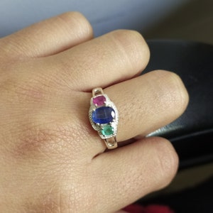 May include: A silver ring featuring three oval-cut gemstones arranged vertically. The top stone is a vibrant pink, the center stone is a deep blue, and the bottom stone is a bright green. The metal band has a textured, beaded edge detail.