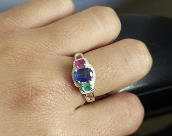 May include: A silver ring featuring three oval-cut gemstones arranged vertically. The top stone is a vibrant pink, the center stone is a deep blue, and the bottom stone is a bright green. The metal band has a textured, beaded edge detail.