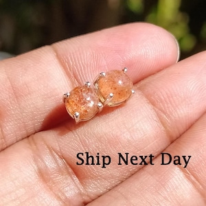 Może przedstawiać: Para srebrnych kolczyków z okrągłymi, pomarańczowymi kamieniami słonecznymi. Kolczyki znajdują się na palcu osoby.