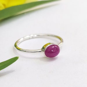 May include: A thin silver ring featuring a single, smooth, oval-shaped magenta gemstone set in a simple metal bezel. The band is polished and slender, showcasing a minimalist design that highlights the vibrant colour of the central stone.