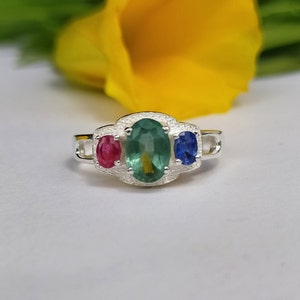 May include: A silver ring with three oval-shaped gemstones. The center stone is a green gemstone, flanked by a red gemstone on the left and a blue gemstone on the right.