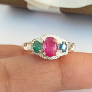 May include: A silver ring with three oval-shaped gemstones. The center stone is a vibrant red ruby, flanked by a green emerald on the left and a blue sapphire on the right.