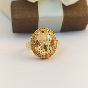 Puede incluir: Un anillo de oro con una gran piedra preciosa ovalada de color amarillo claro. La piedra preciosa está rodeada por un borde texturizado y con cuentas. El anillo está engastado en una banda de oro. El fondo incluye una caja de regalo marrón con una cinta blanca.