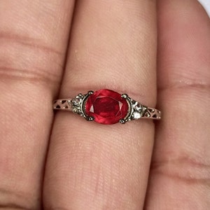 May include: A silver ring with a red oval gemstone and two smaller clear gemstones on either side.