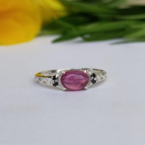 Könnte beinhalten: Ein silberner Ring mit einem rosafarbenen ovalen Edelstein und zwei schwarzen Edelsteinen auf jeder Seite.