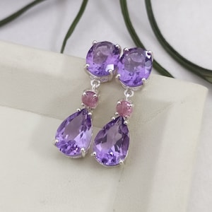 May include: A pair of dangle earrings with amethyst and pink stones. The earrings feature a round amethyst stone at the top, followed by a smaller round pink stone, and a teardrop-shaped amethyst stone at the bottom.