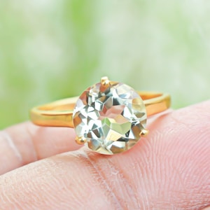 Könnte beinhalten: Ein goldener Ring mit einem großen, runden, facettierten, hellgrünen Edelstein. Der Ring hat ein goldenes Band und goldene Krappen. Der Hintergrund ist ein weiches, verschwommenes Grün.